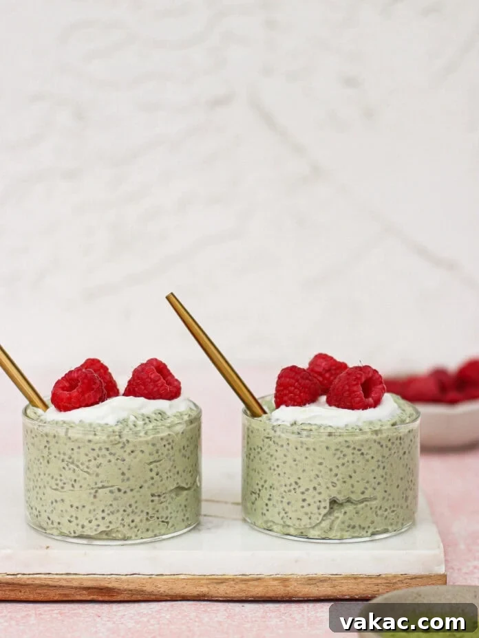 Quick High Protein Matcha Chia Pudding with Yogurt 6 Two glass cups filled with green chia pudding, topped with white yogurt and fresh raspberries, each with a gold straw, sit on a marble board. A bowl of raspberries and a dish of matcha powder are nearby.