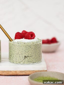 A small glass of matcha chia pudding topped with whipped cream and fresh raspberries sits on a white tray. A gold spoon is inside the glass. Bowls with raspberries and green powder are nearby against a light pink background.
