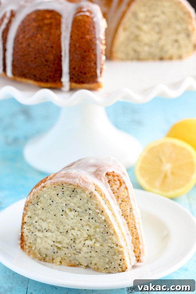 Zesty Lemon Poppy Seed Bundt Cake 2 A beautifully sliced, glazed lemon poppy seed bundt cake resting on a pristine white plate, with the larger cake on an elegant stand in the background. The image highlights the cake's moist interior, golden hue, and the glossy lemon glaze.
