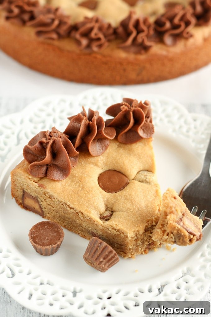 A slice of homemade cookie cake on a white plate with a fork. The rest of the cookie cake rests in the background, showing its soft texture and generous frosting.