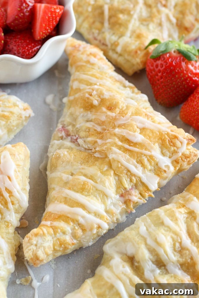 Flaky Strawberry Cream Cheese Pockets 3 Several freshly baked strawberry cream cheese turnovers laid out on a sheet of parchment paper. A white bowl brimming with sliced fresh strawberries sits nearby, highlighting the key ingredient used in these tempting pastries.