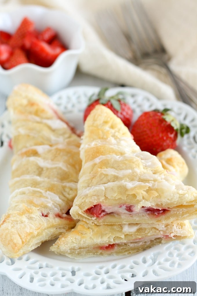 Flaky Strawberry Cream Cheese Pockets 4 Two freshly baked strawberry cream cheese turnovers presented on a pristine white plate. One turnover has been artfully cut in half and stacked to showcase its inviting layers of flaky pastry, bright red strawberries, and creamy white cheese filling. Fresh strawberries and elegant forks adorn the background.