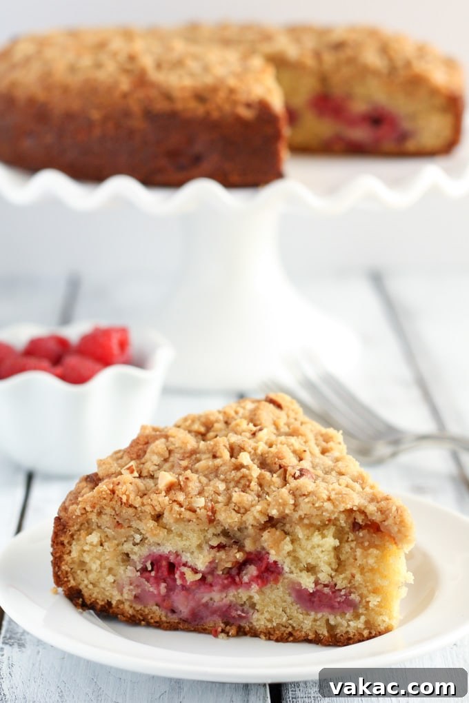 Blissful Raspberry Almond Cake 2 A beautiful slice of raspberry almond cake with a generous crumb topping, presented elegantly on a white plate. In the blurred background, a bowl of fresh, plump raspberries and a full cake on a stand hint at the bounty of this delicious creation.