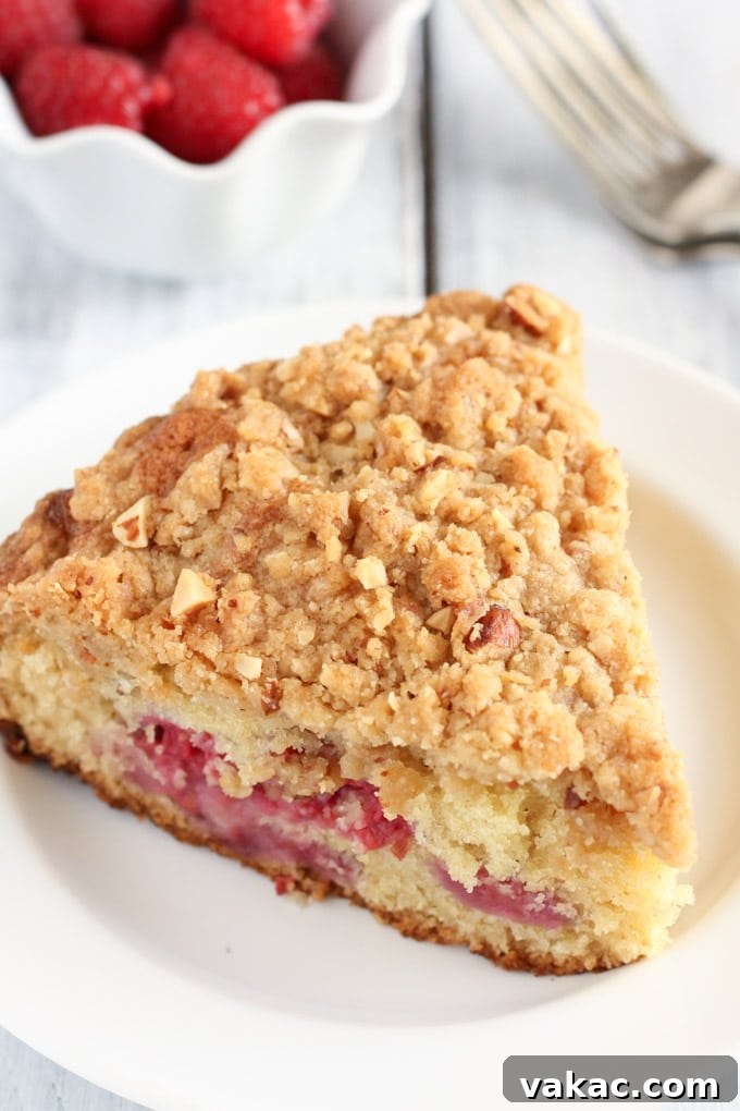 Blissful Raspberry Almond Cake 4 A pristine slice of raspberry almond cake with a golden crumb topping rests on a white plate, ready to be enjoyed. In the background, a small bowl of fresh, ripe raspberries awaits, emphasizing the key fruit in this delightful dessert.