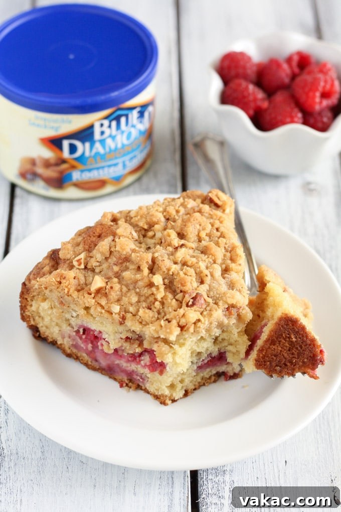 Blissful Raspberry Almond Cake 5 A perfectly baked slice of raspberry crumb cake, topped with golden almonds, sits on a clean white plate. The background features a bowl of fresh, bright raspberries and a container of delicious Blue Diamond Roasted Salted Almonds, highlighting the key components of this delightful dessert.