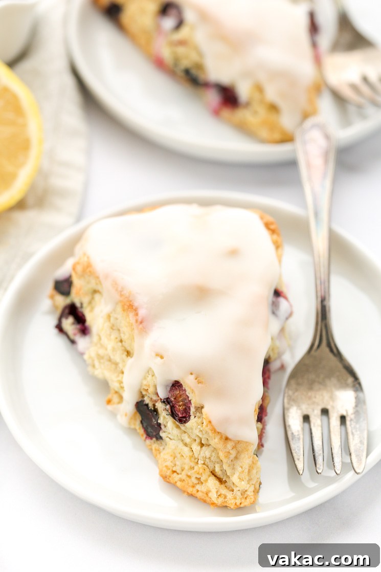 Bright Lemon Blueberry Scones 2 A perfectly baked lemon blueberry scone, glistening with lemon glaze, rests on a pristine white plate. In the soft-focus background, another scone and fresh lemon slices hint at the delightful flavors within this homemade pastry.
