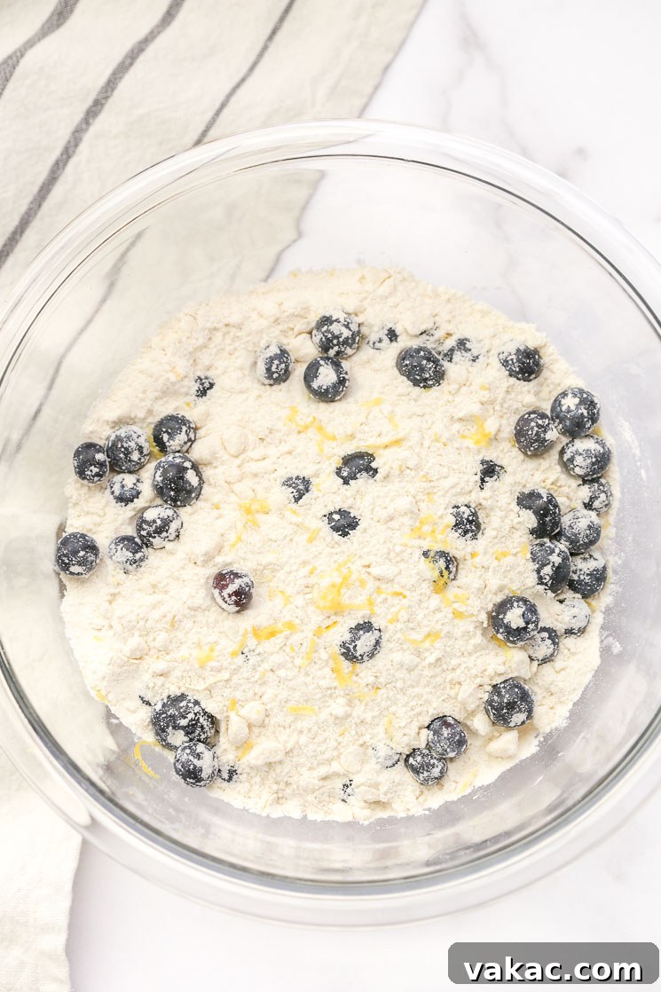 Bright Lemon Blueberry Scones 3 A clear glass bowl showcasing the perfectly combined dry ingredients, vibrant fresh blueberries, and aromatic lemon zest, ready for the wet ingredients. A light gray napkin rests elegantly beside the bowl.