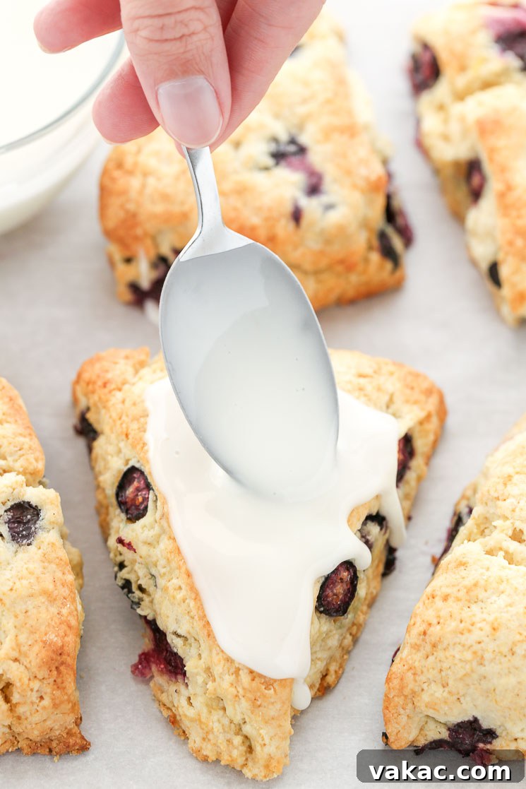 Bright Lemon Blueberry Scones 5 A mouthwatering close-up captures the moment of pure delight as fresh lemon glaze is delicately drizzled over warm, golden-brown lemon blueberry scones resting on parchment paper. The vibrant berries peek out from the tender crumb, promising an explosion of flavor.