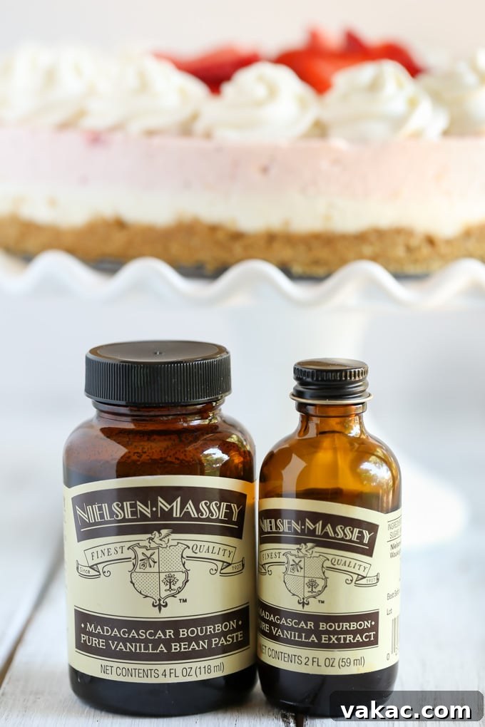 Velvet Strawberry Vanilla Bean Cheesecake 4 A close-up view of two distinct bottles: one of Nielsen-Massey vanilla bean paste and another of vanilla extract. Behind them, slightly blurred, sits a fresh strawberry cheesecake on an elegant cake stand, highlighting the key ingredients used in its creation.