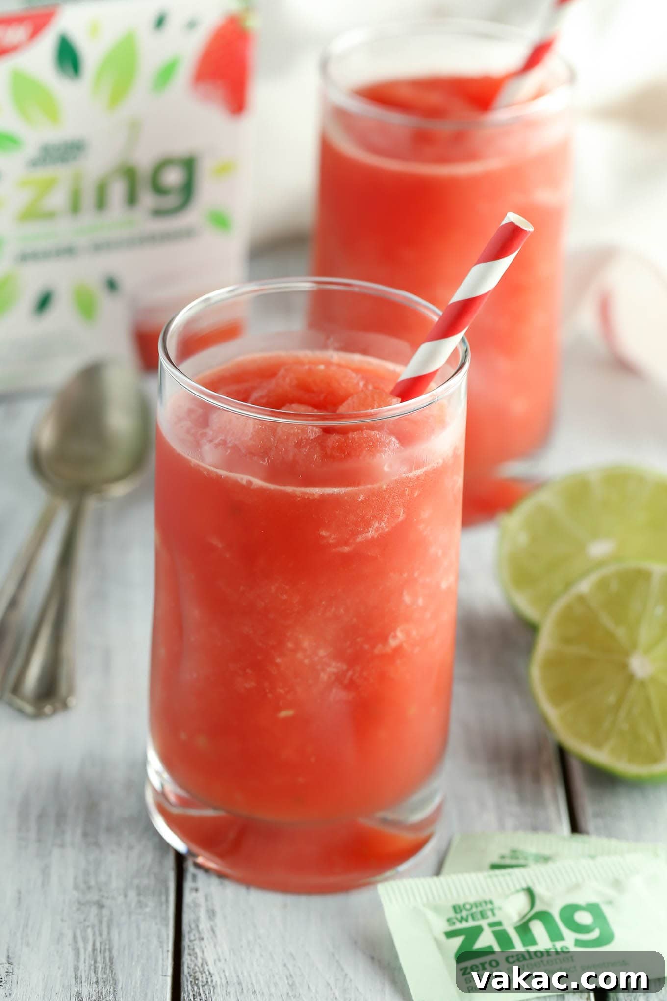 Zesty Watermelon Freeze 2 Two glasses of refreshing pink watermelon lime slushies garnished with fresh lime wedges, sitting on a light wooden surface with sliced watermelon and limes nearby.