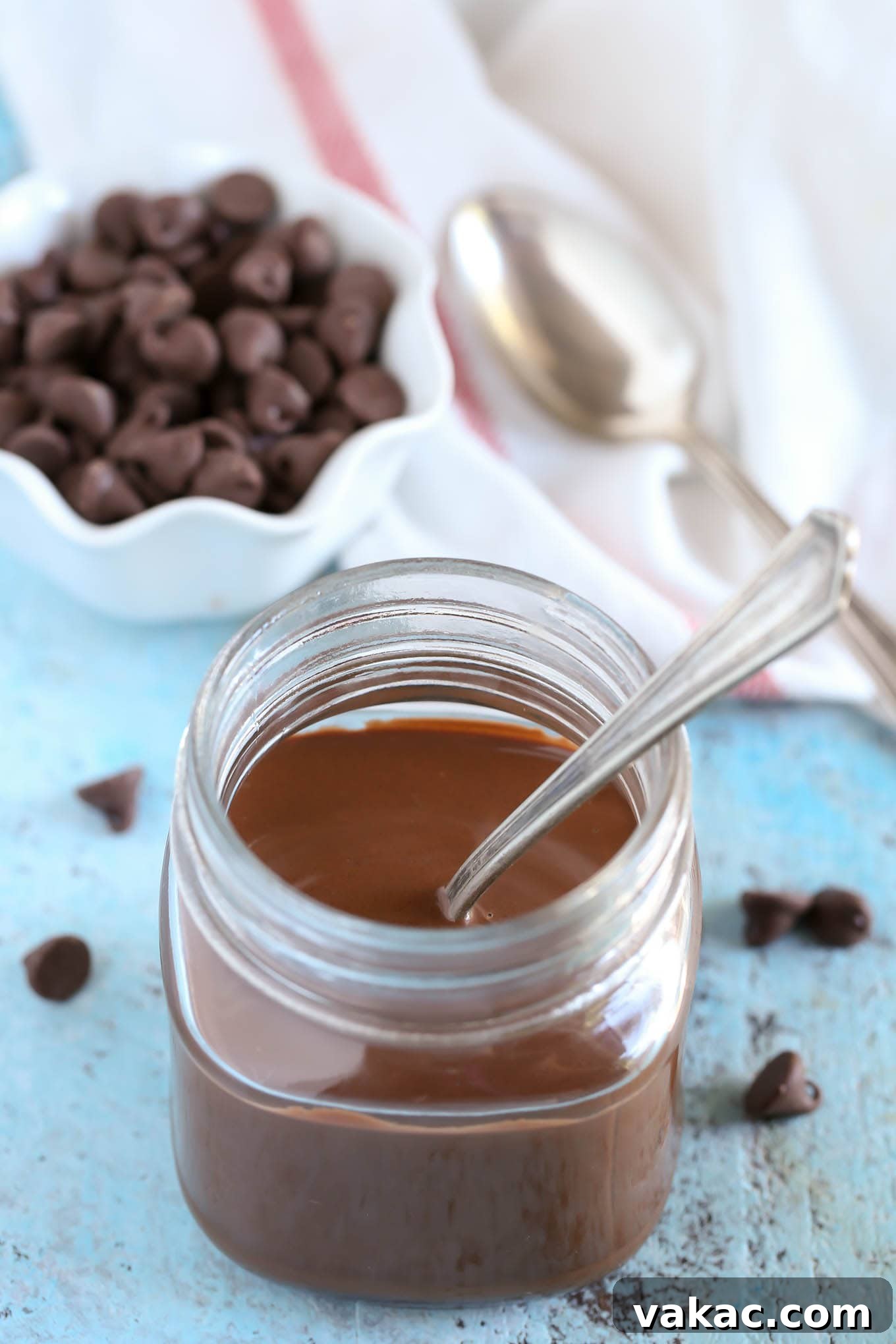 DIY Chocolate Crunch Topping 2 Homemade magic shell in a glass jar. A spoon is sticking out of the jar. A small dish of chocolate chips, a tea towel, and a spoon rest in the background.