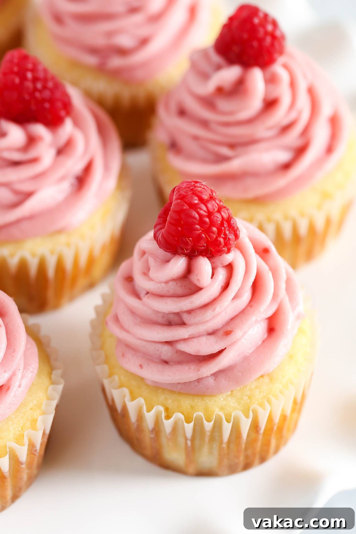 Lemon Raspberry Bliss Cupcakes 2 Close-up of a perfectly frosted lemon cupcake topped with fresh raspberries, set on a white platter amidst other cupcakes.