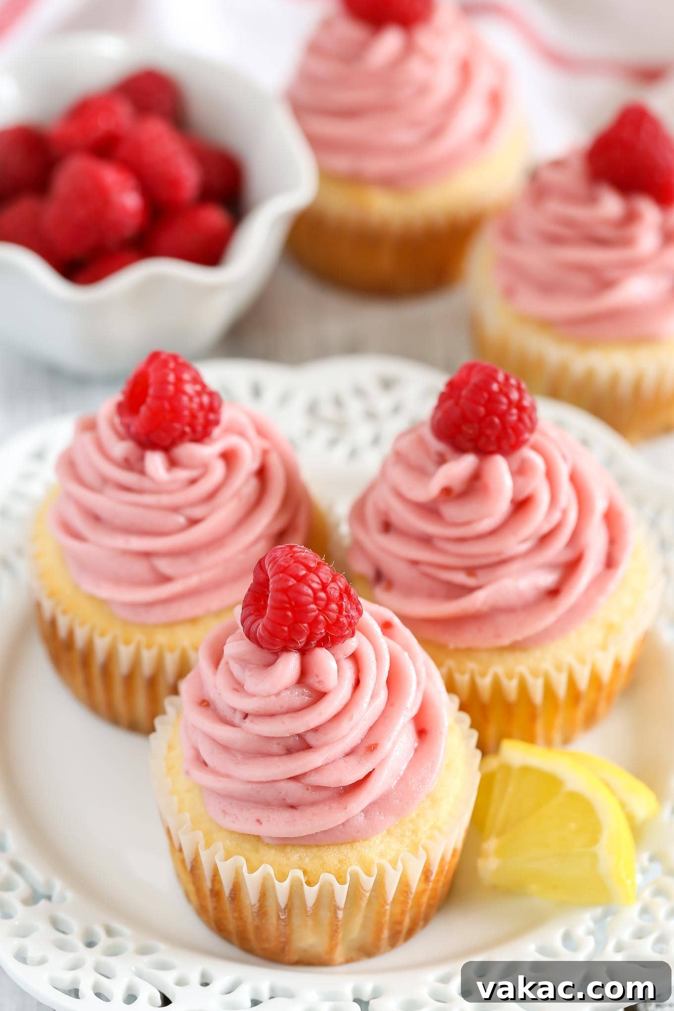Lemon Raspberry Bliss Cupcakes 3 Collection of lemon cupcakes adorned with raspberry frosting and fresh raspberries on a pristine white plate, with a bowl of berries and more cupcakes artfully arranged in the background.