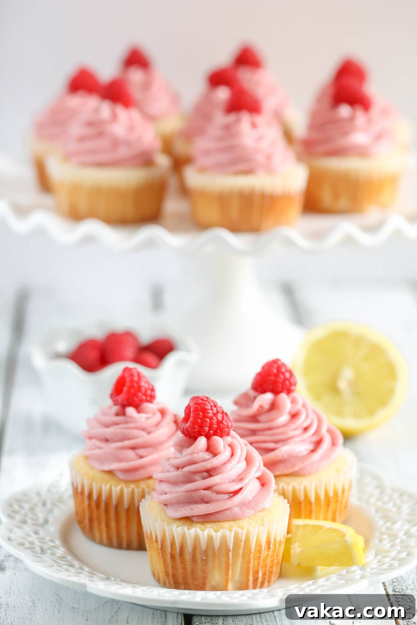 Lemon Raspberry Bliss Cupcakes 4 Lemon cupcakes topped with fluffy raspberry frosting and garnished with fresh raspberries, displayed on a white plate with more cupcakes on a cake stand in the background.