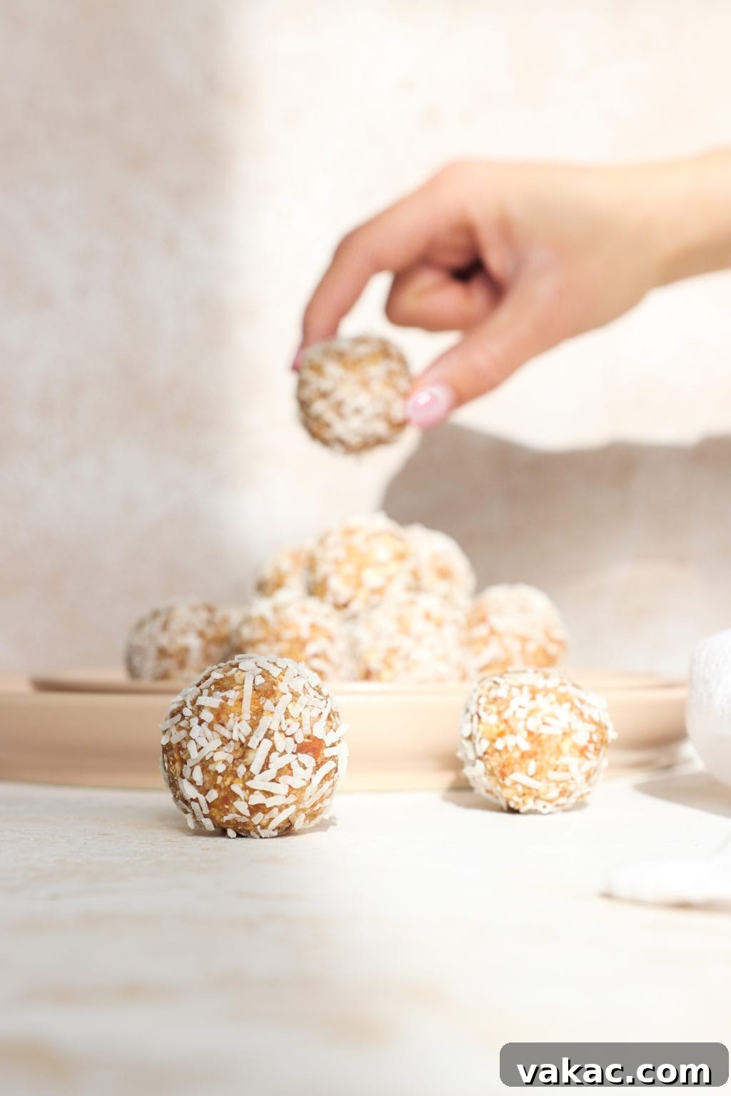 Two freshly prepared coconut lemon energy balls are in the foreground, beautifully placed, while a hand reaches into the soft-focus background to pick up another energy ball from a plate, emphasizing the irresistible appeal of these healthy snacks.