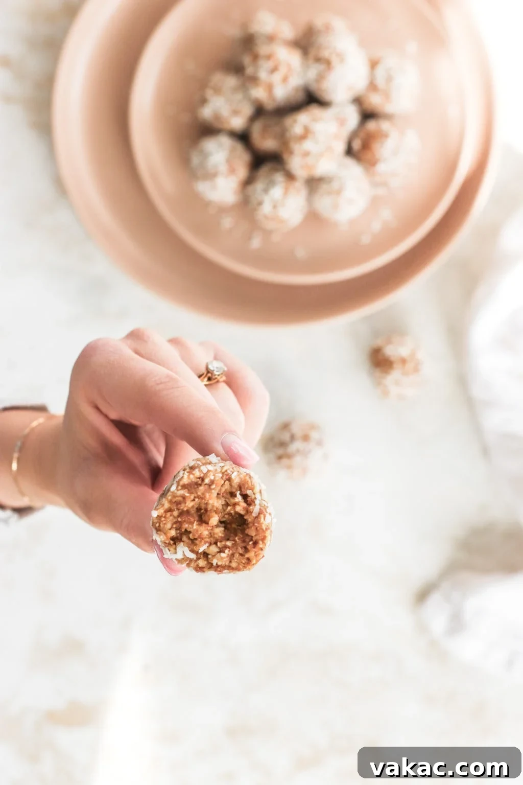 A close-up shot of a hand holding a lemon coconut energy ball, with a small bite taken out, revealing the wholesome texture inside. In the blurred background, a plate full of more energy balls awaits, highlighting their delicious and ready-to-enjoy nature.