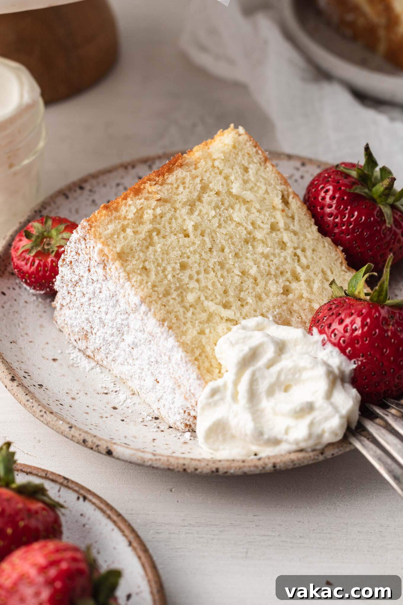 A perfectly sliced piece of chiffon cake on a white speckled plate, garnished with fresh strawberries and a cloud of whipped cream, showcasing its light and airy texture.
