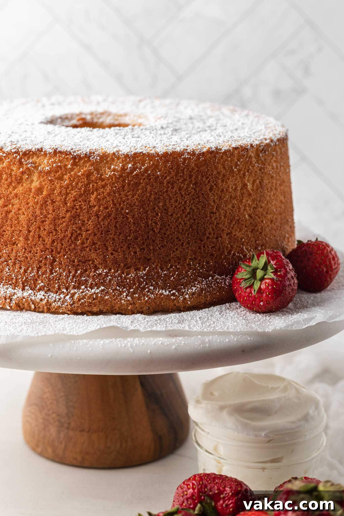 A freshly baked chiffon cake, beautifully dusted with powdered sugar, resting on a marble cake stand alongside fresh strawberries.