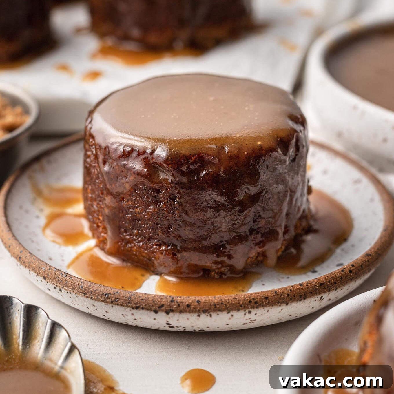 A small round date cake covered in toffee sauce on a white speckled plate.