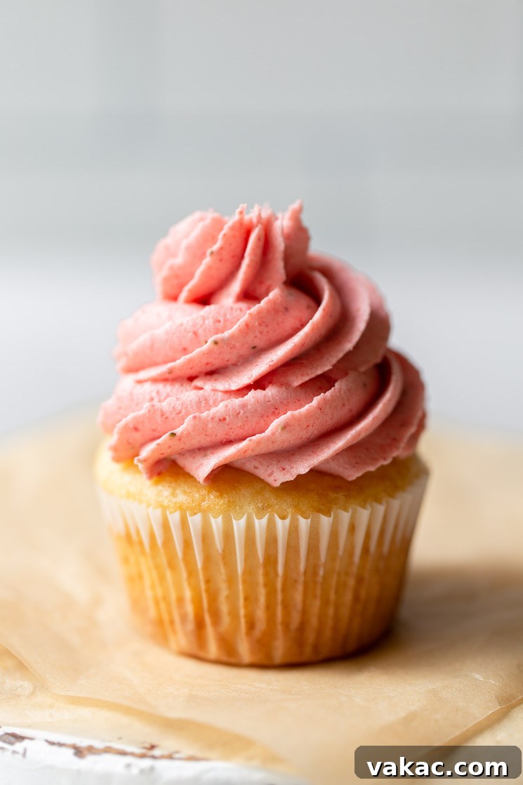 A single, beautifully frosted cupcake sits delicately on a piece of brown parchment paper, adorned with a swirl of vibrant strawberry buttercream frosting.