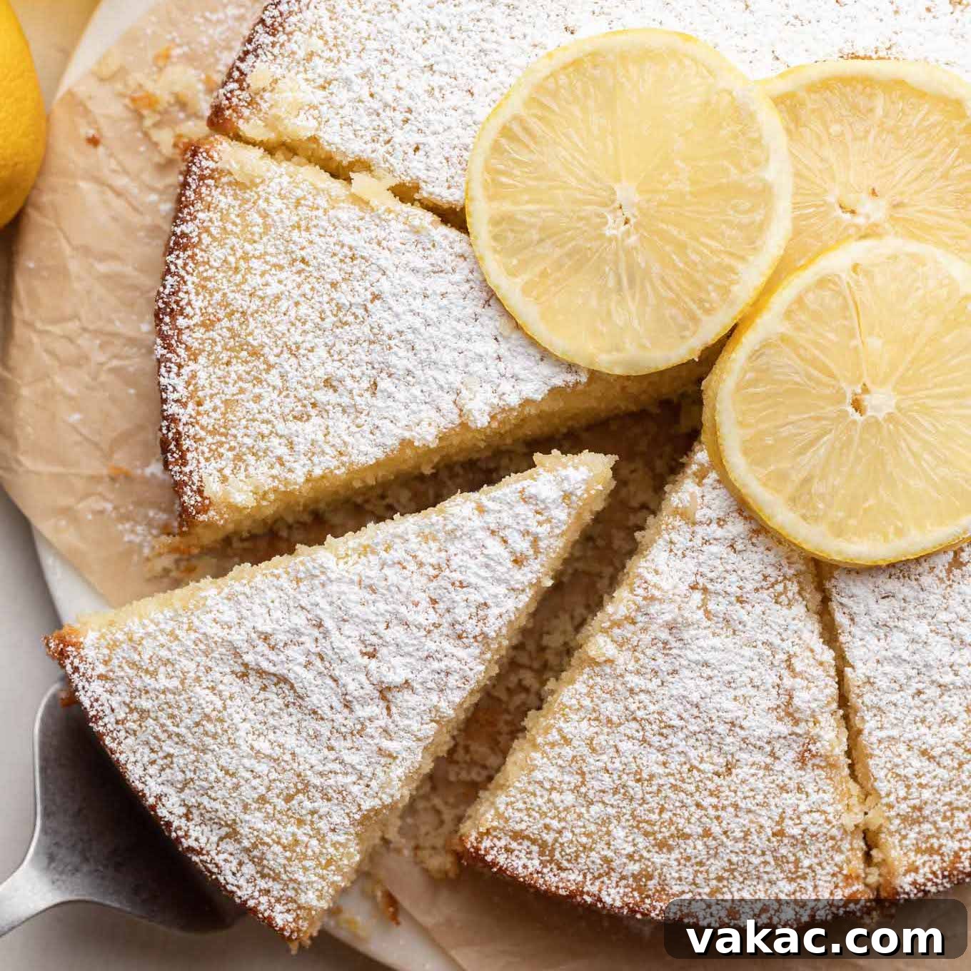 A beautifully baked and sliced lemon olive oil cake on a piece of parchment paper. One slice is being gently removed, ready to be served.