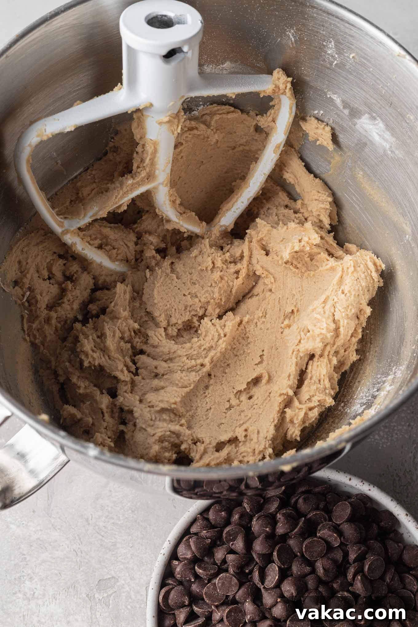 Egg-Free Chocolate Chip Bliss 5 No-egg cookie dough in a metal mixing bowl with a paddle attachment. A bowl of chocolate chips rest on the side.