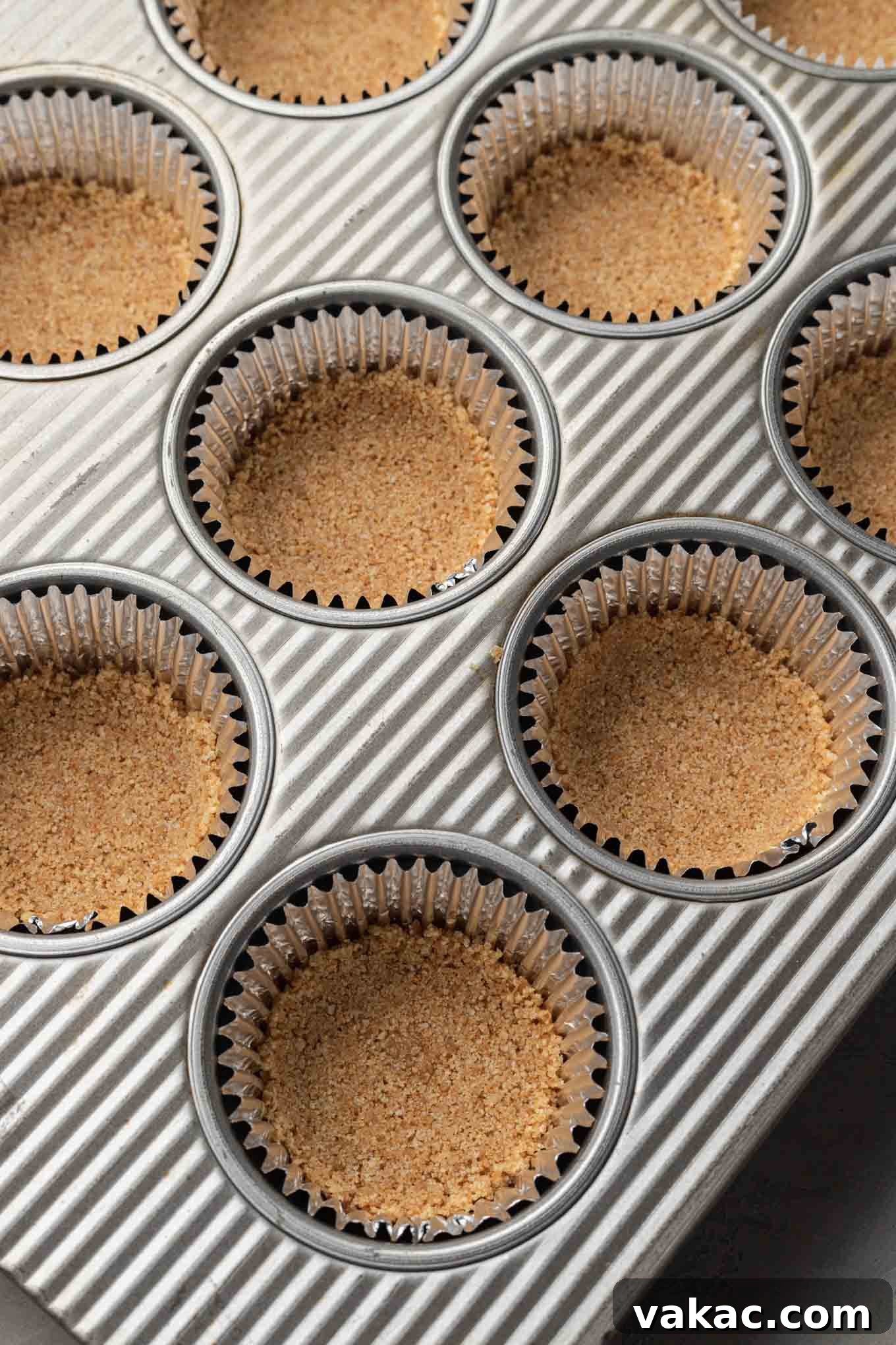 Effortless Mini Cheesecake Delights 4 Muffin cups meticulously lined with cupcake liners and uniformly filled with a pressed graham cracker crust.