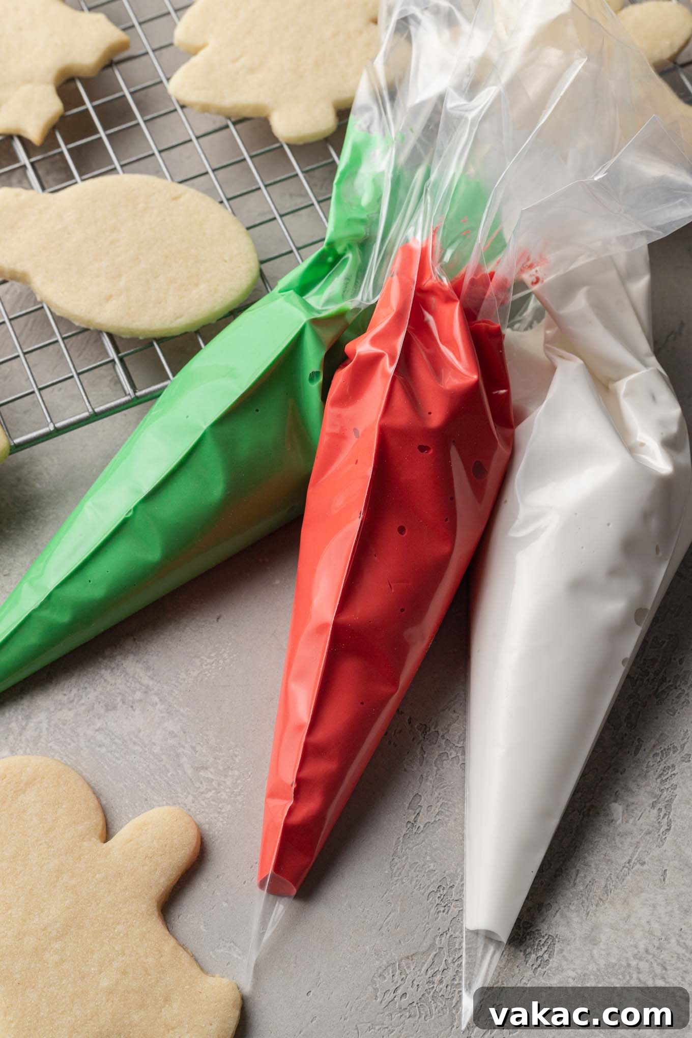 An overhead view of three piping bags filled with red, green, and white royal icing. A few sugar cookies are laying on a cooling rack by the piping bags.