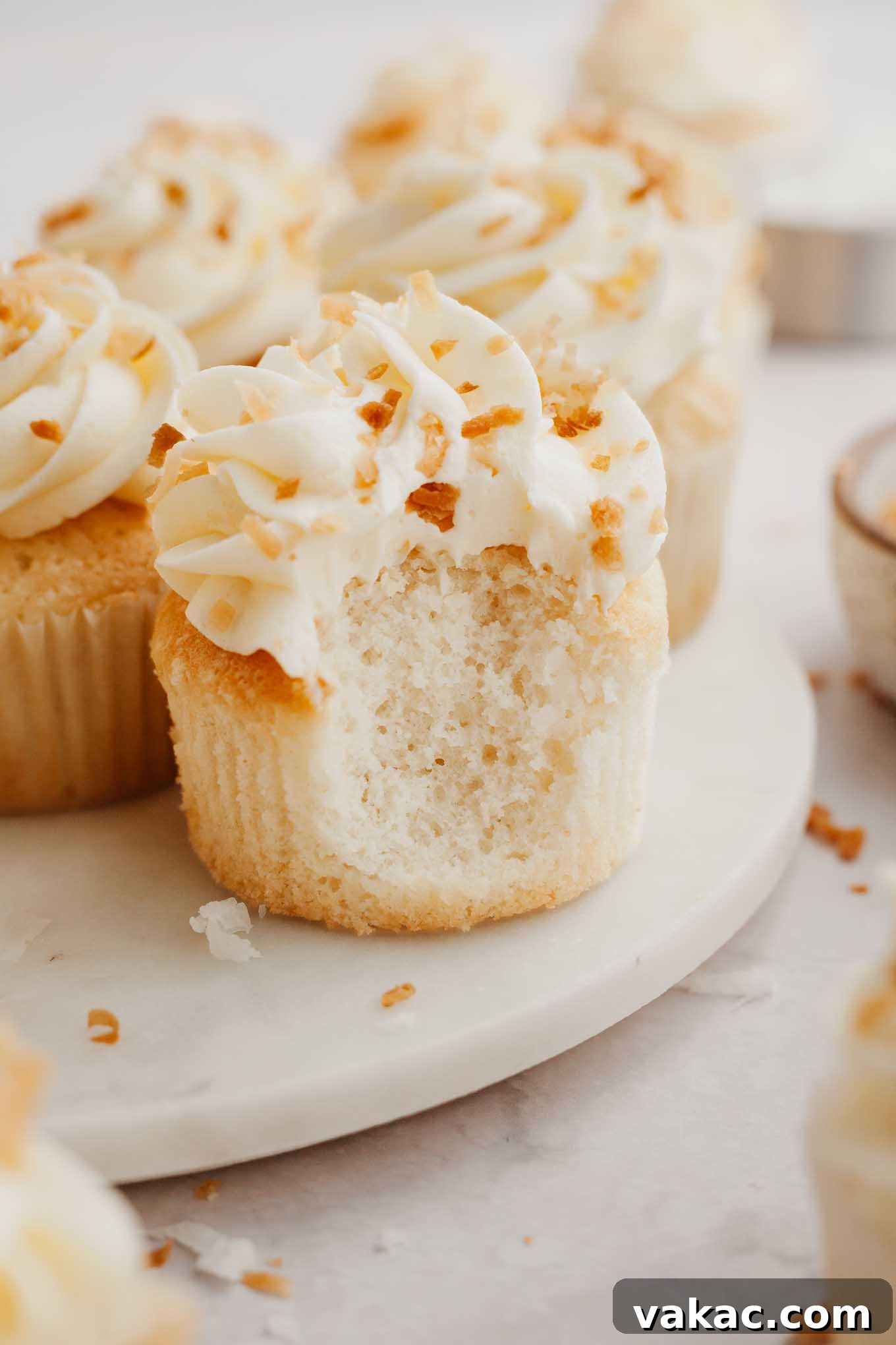 Tropical Coconut Delights 11 A delicious coconut cupcake with a small bite taken out, revealing its moist interior, placed elegantly on a plate with more cupcakes in the background.