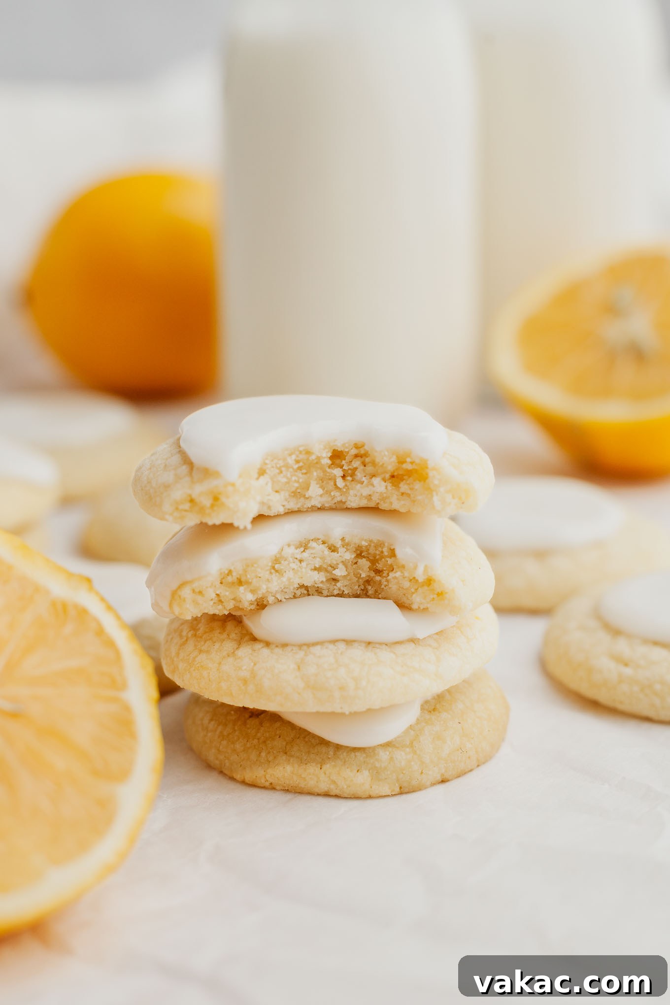 A stack of four golden lemon meltaway cookies, glistening with a fresh lemon glaze, surrounded by more cookies on a rustic background.