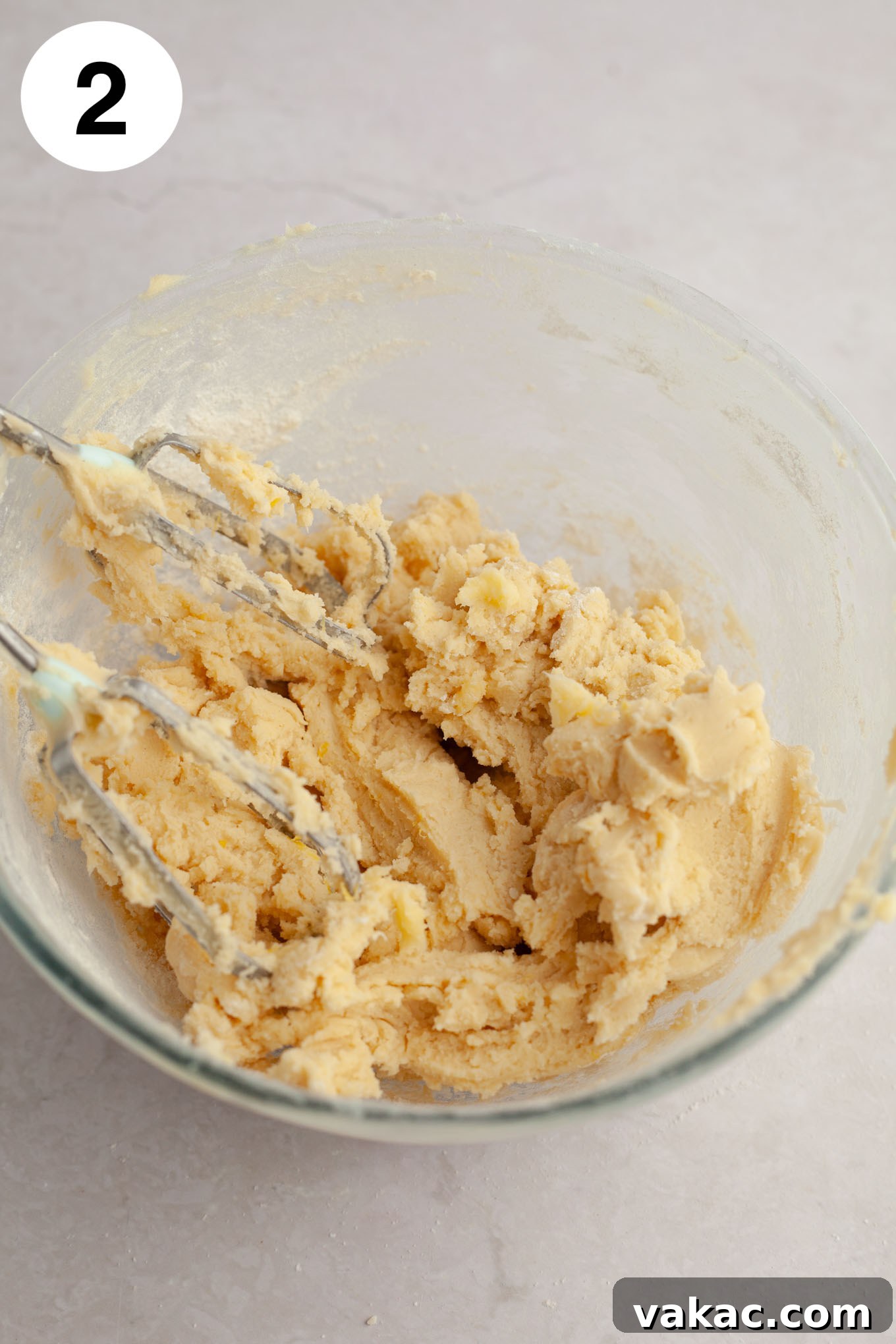 An overhead view of light yellow lemon cookie dough in a glass mixing bowl, with electric beaters resting nearby, showing the smooth, cohesive texture.