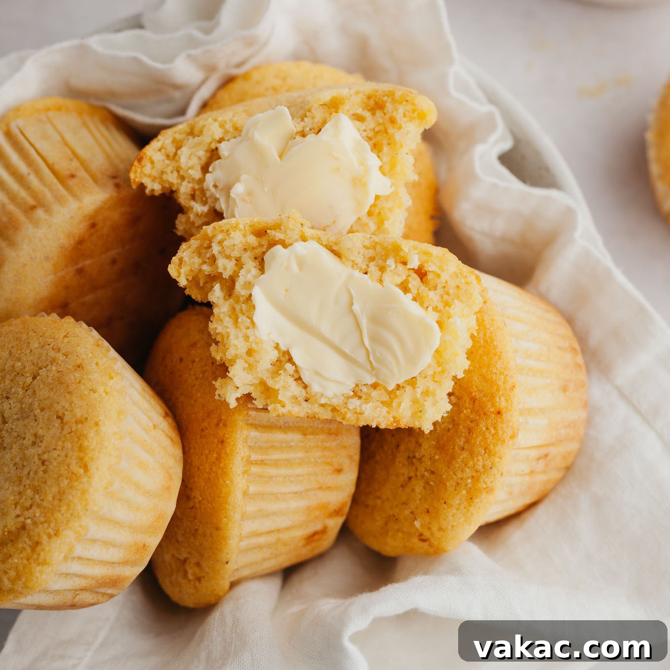 Golden Fluffy Cornbread Muffins 8 A close-up of a pile of cornbread muffins in a towel-lined serving bowl. The top muffin is split in half, revealing its tender crumb and slathered with melting butter.
