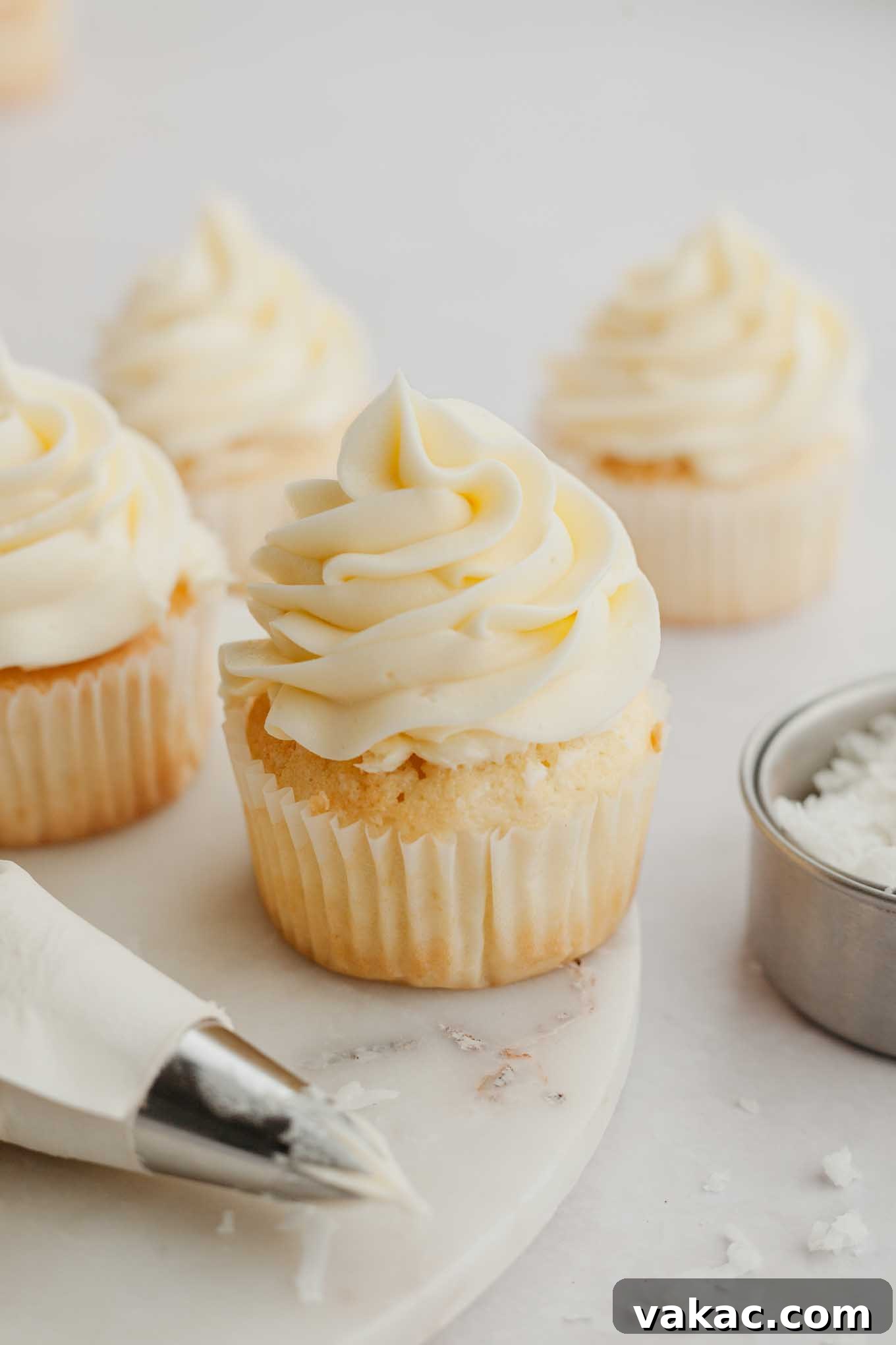 A close-up shot of several fluffy cupcakes generously topped with creamy white coconut frosting, garnished with delicate toasted coconut flakes.