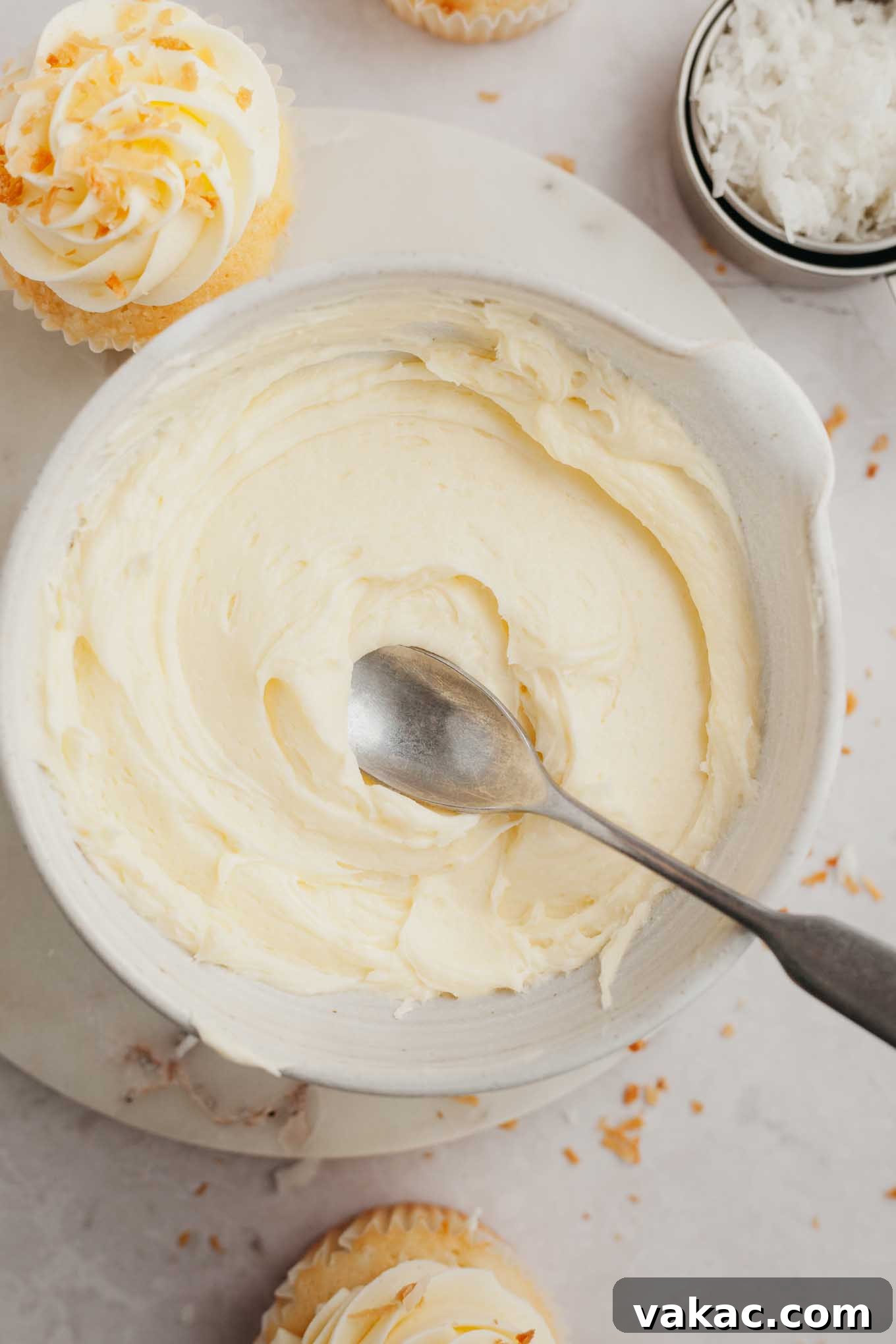 A ceramic bowl brimming with fluffy, white coconut frosting, with a silver spoon gently resting inside, inviting a taste.