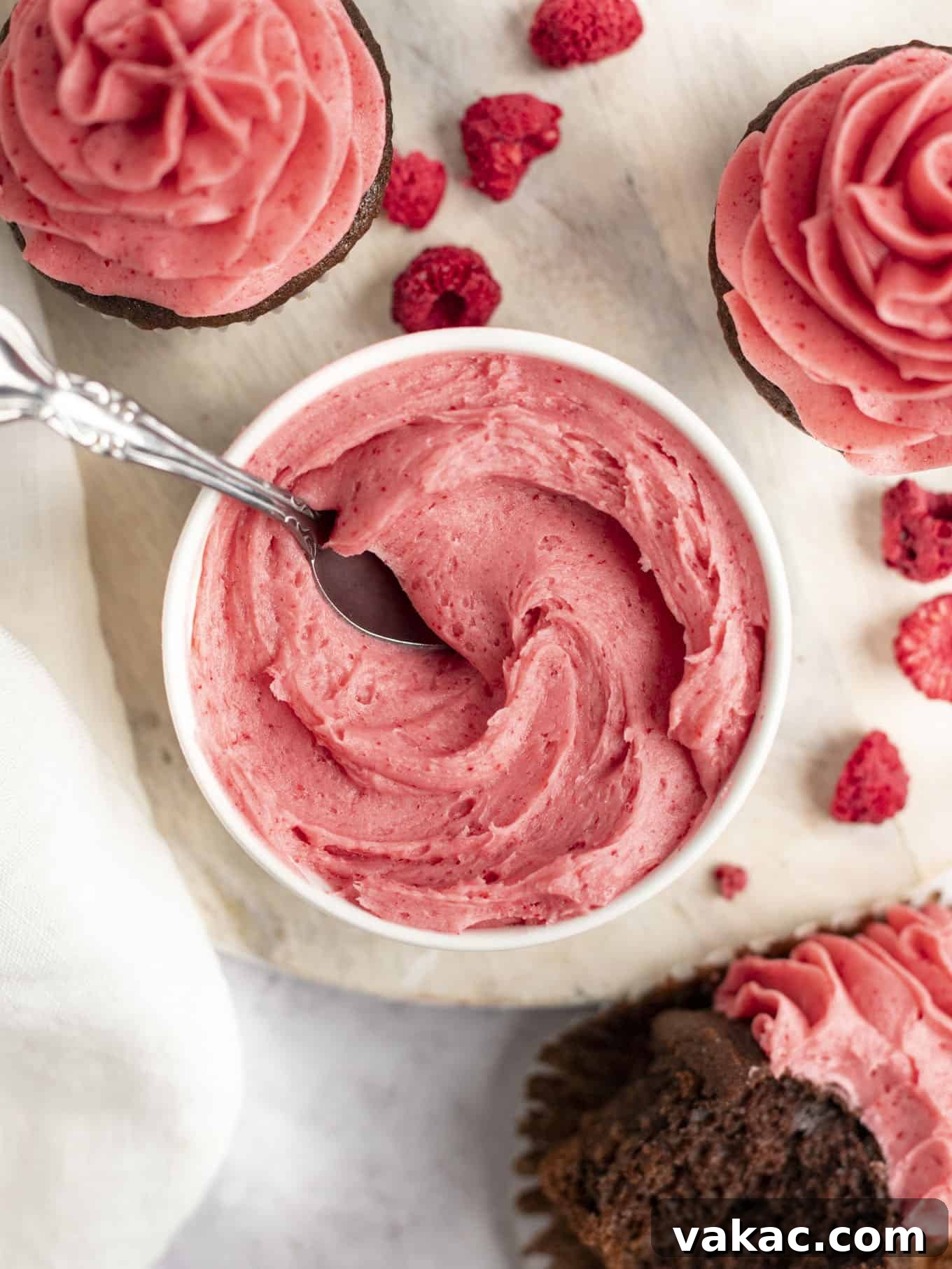 Velvet Raspberry Icing 16 An overhead view of smooth, pale pink raspberry buttercream in a white ceramic bowl, with a silver spoon sticking out, inviting a taste.