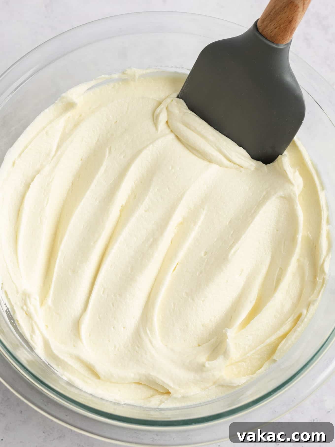 Velvet White Chocolate Frosting 5 A large bowl of smooth, white chocolate frosting with a rubber spatula resting on the side, ready for use.