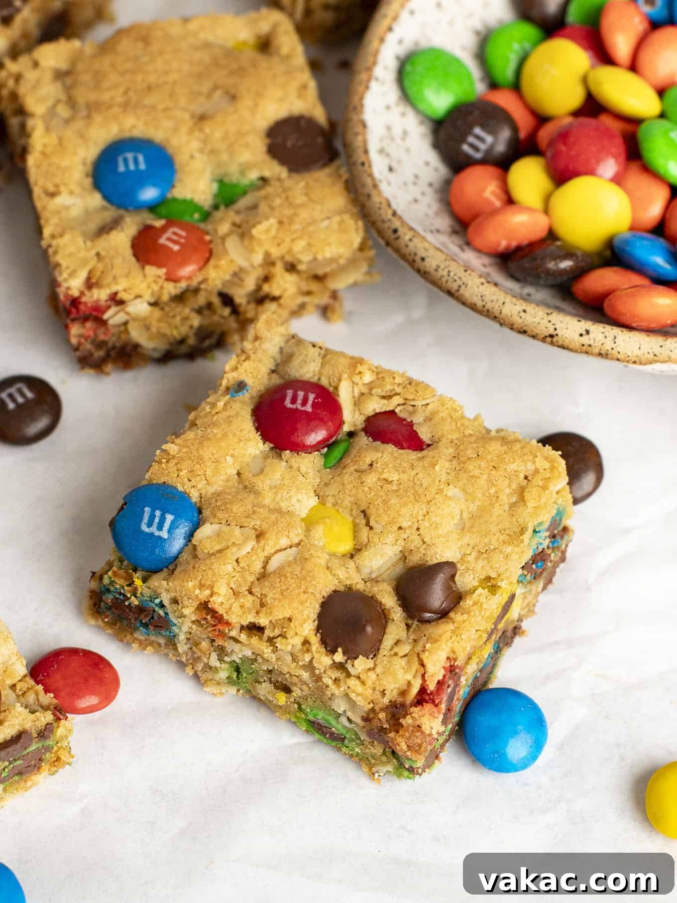 Ultimate Everything Cookie Bars 11 A close-up of a single monster cookie bar, showcasing its soft interior, visible oats, and abundant M&M's and chocolate chips, next to a small dish of M&M's.
