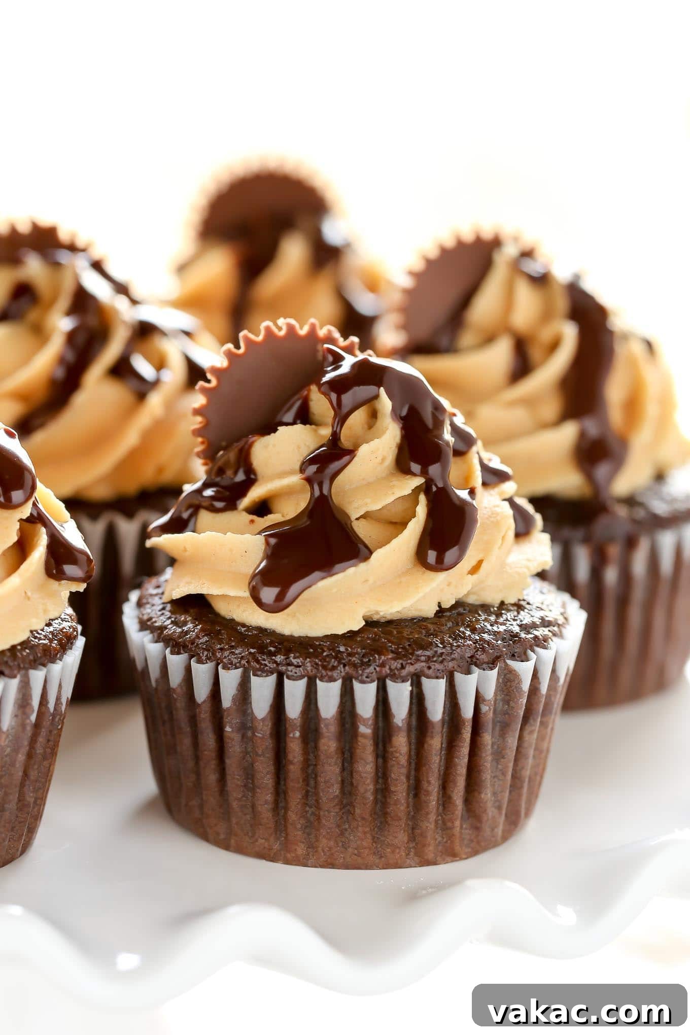 Peanut Butter Cup Chocolate Cupcakes 2 Close up of chocolate peanut butter cupcakes on a white cake stand, showcasing the creamy frosting, chocolate ganache drizzle, and peanut butter cup topping.