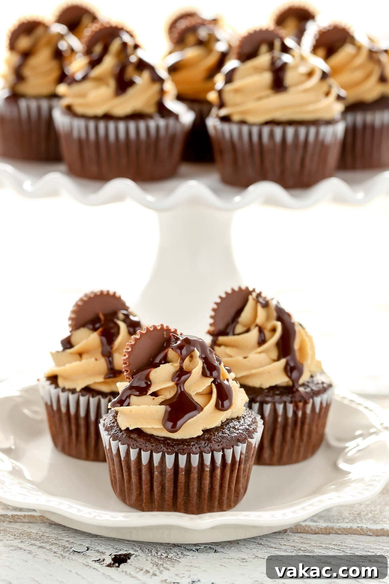 Peanut Butter Cup Chocolate Cupcakes 4 Three meticulously decorated chocolate peanut butter cupcakes displayed on a white plate, with a cake stand full of additional cupcakes blurred in the background.