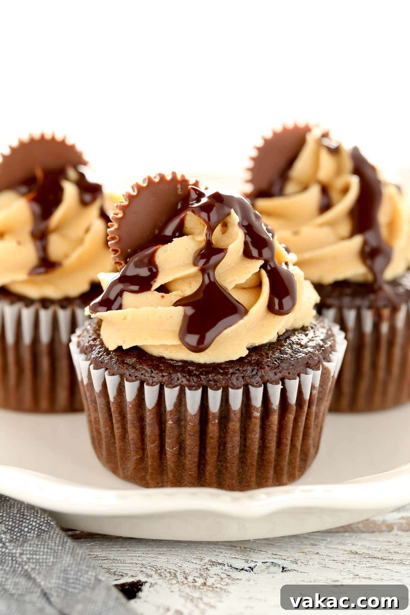 Peanut Butter Cup Chocolate Cupcakes 5 A close-up shot of three decorated chocolate peanut butter cupcakes on a white plate, highlighting the detail of the frosting, ganache, and peanut butter cup toppings.