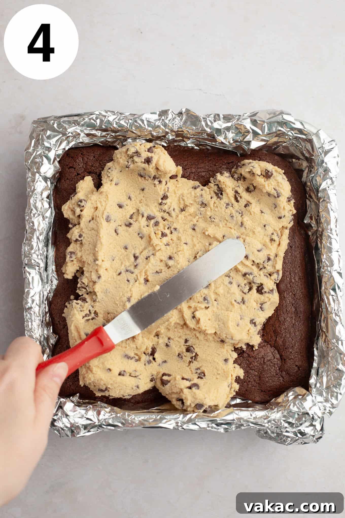 Brownie Cookie Dough Perfection 7 An overhead view of edible chocolate chip cookie dough being carefully spread over a cooled, rectangular pan of brownies, creating an even layer.