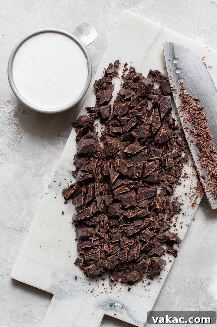 A clean marble cutting board displaying neatly chopped semi-sweet chocolate alongside a small measuring cup filled with rich heavy cream.