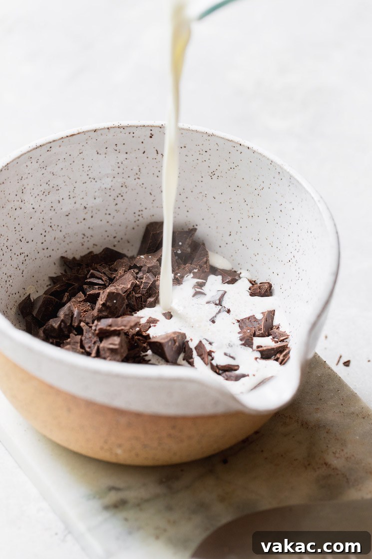 A small bowl holding finely chopped chocolate, with warm heavy cream being gently poured over the top, initiating the ganache process.