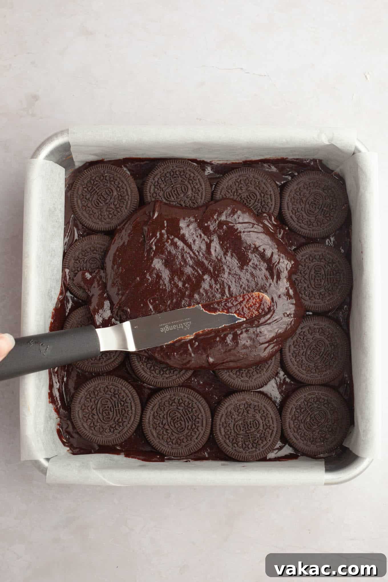 An overhead view of the remaining brownie batter being carefully spread and smoothed over the layer of whole Oreos in the baking pan, ensuring complete coverage.