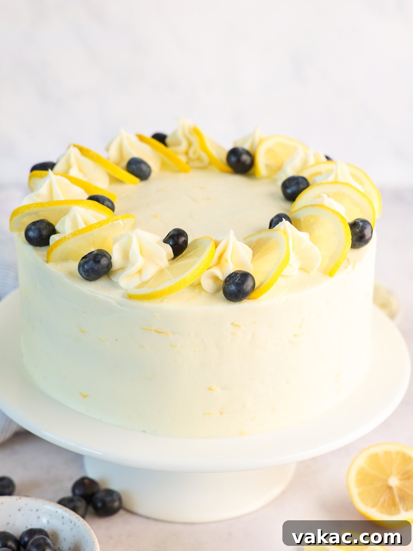 Sunshine Berry Cloud Cake 2 A stunning three-layer lemon blueberry cake with lemon cream cheese frosting on a decorative cake stand, garnished with fresh blueberries and lemon slices.