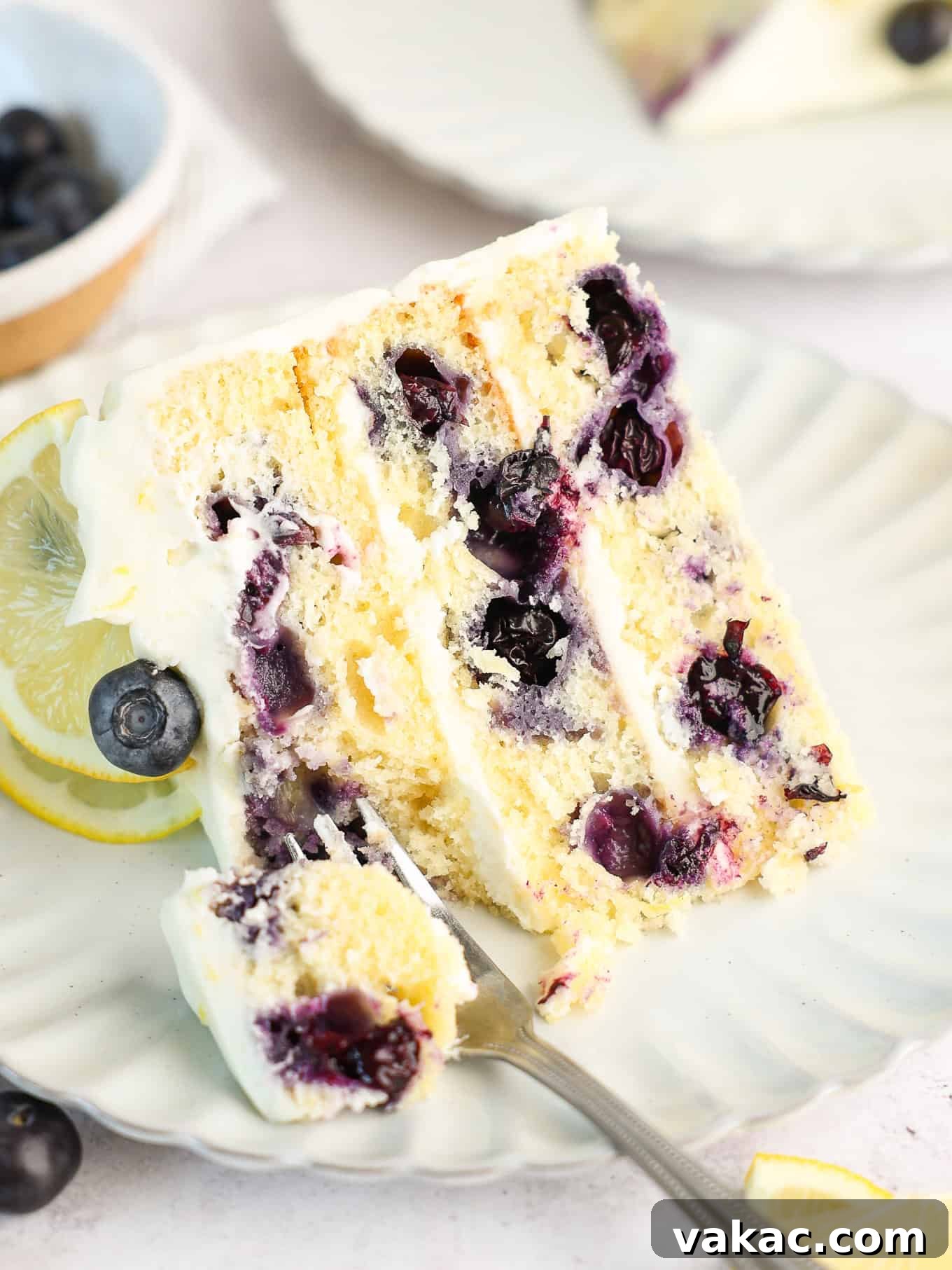Sunshine Berry Cloud Cake 14 A beautiful slice of lemon cake with visible blueberries, served on a white dessert plate with a fork.