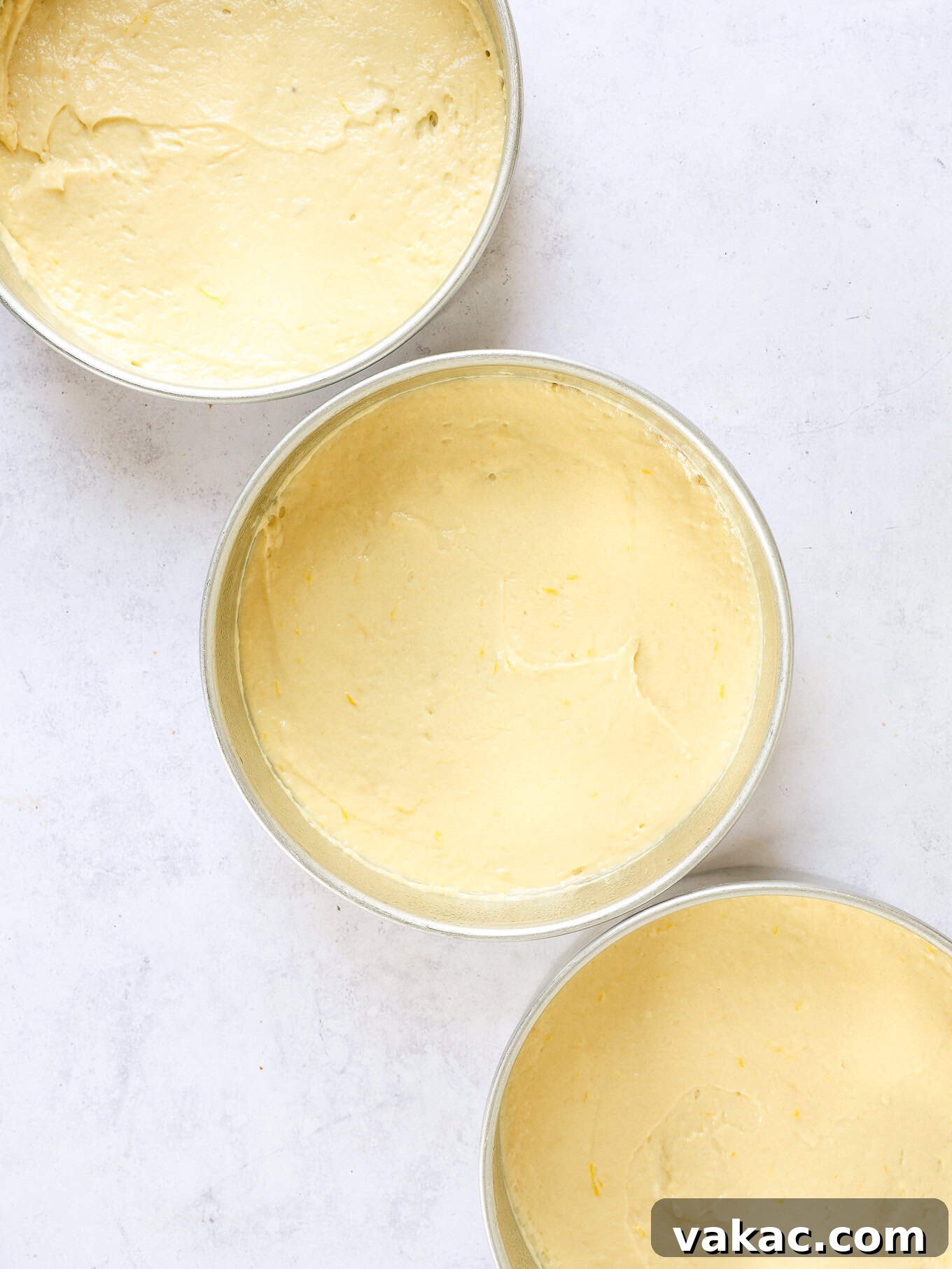 Sunshine Berry Cloud Cake 7 Three empty round cake pans, each lined with parchment paper at the bottom, with a small amount of plain lemon cake batter spread thinly in each.