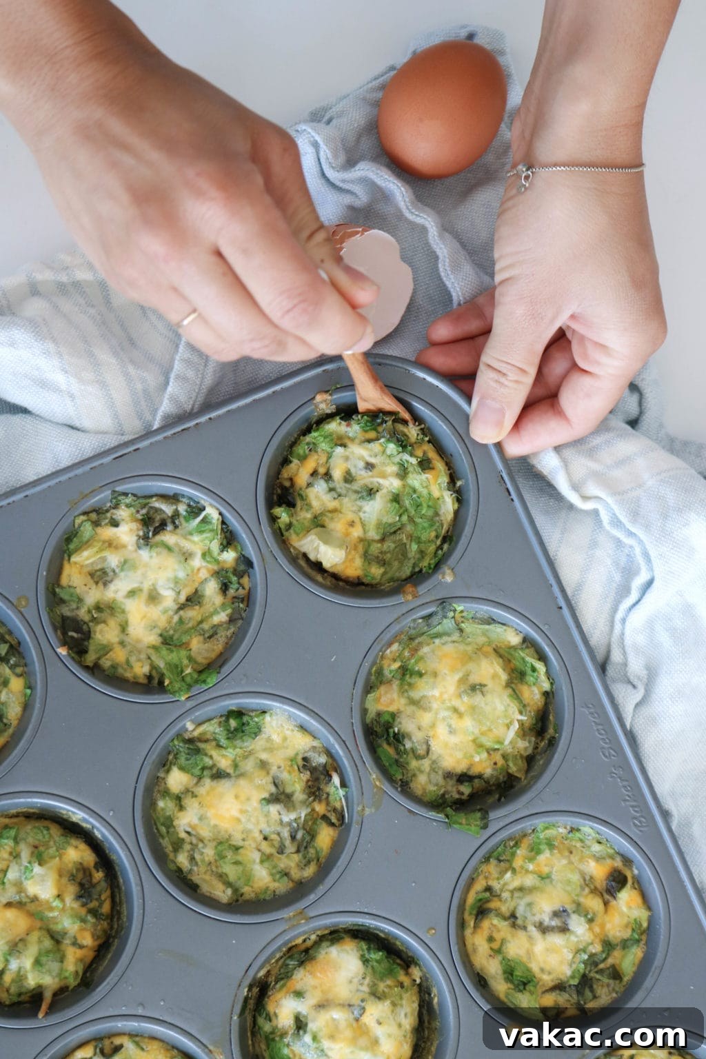 Easy Meal Prep Egg Muffins 2 Muffin tin frittatas with eggs, cheese, and mushrooms are in a muffin tin, laid over a blue tea towel. Cracked egg shells are on the side and a woman's hand is scooping one of the frittatas out with a small wooden spoon.