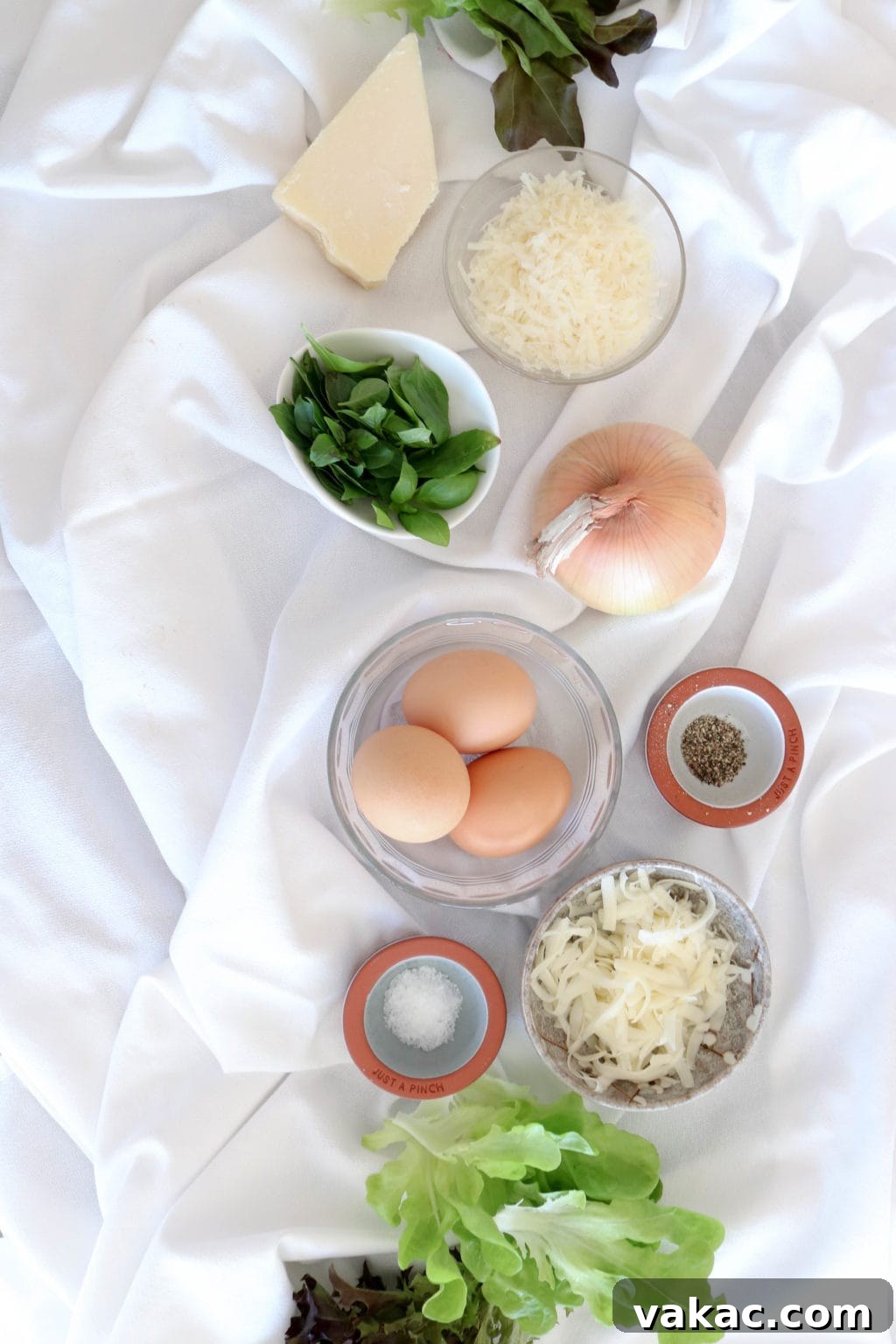 Easy Meal Prep Egg Muffins 3 Ingredients for muffin tin frattatas are laid out on a white wrinkled tablecloth and include inspired greens lettuce, parmesan cheese, basil, onion, eggs, salt and pepper.