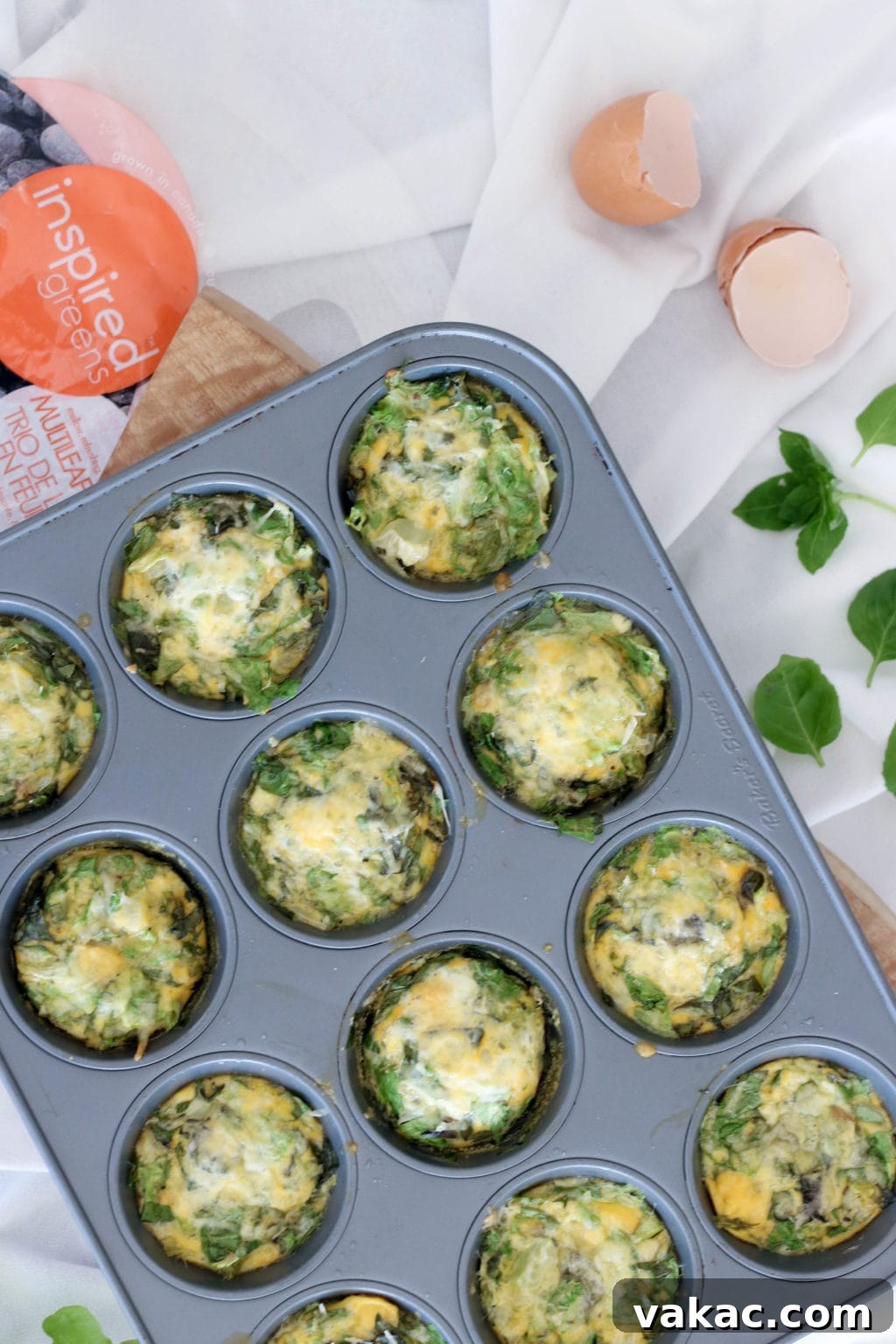 Easy Meal Prep Egg Muffins 4 Muffin tin frittatas in a medium sized muffin tin over a white sheet with a bag of inspired greens beside it and a cracked egg. Ingredients include eggs, butter, greens, onion, basil, cheese.